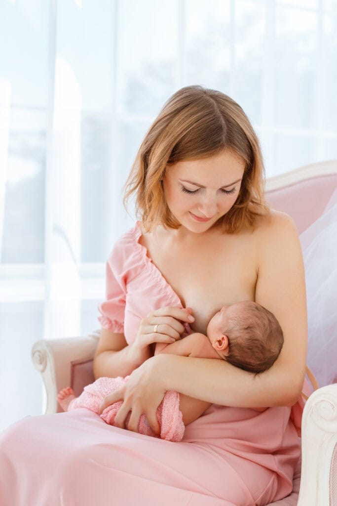 A mother in a soft pink dress breastfeeds her newborn in a calm, sunlit room—highlighting the nurturing care and close observation often involved in tongue tie evaluation and feeding support.