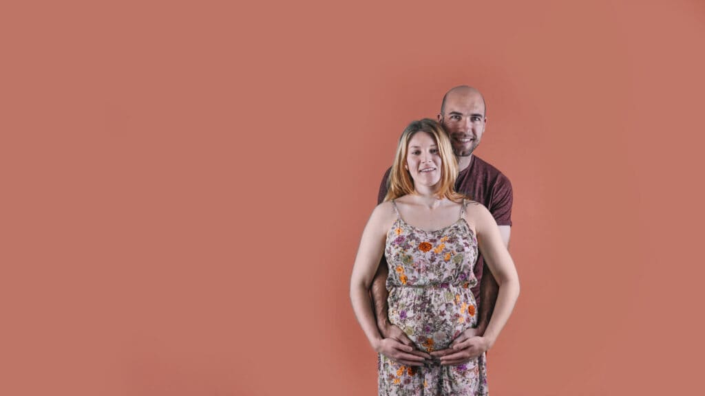 A smiling pregnant woman stands with her partner behind her, both cradling her belly—symbolizing preparation, support, and confidence as they get ready for breastfeeding and parenthood together.