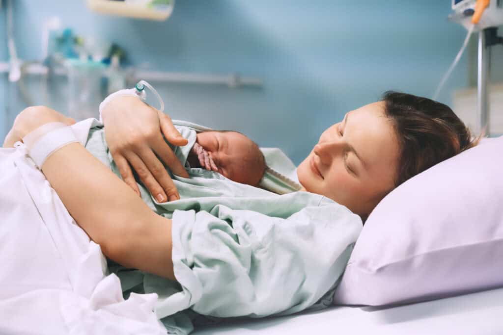A mother lies in a hospital bed holding her premature newborn skin-to-skin, resting peacefully together after NICU care—symbolizing the early bonding and first steps toward breastfeeding after medical separation.
